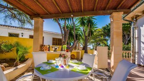 A covered outdoor dining area with a table set for two, surrounded by white chairs, under a wooden pergola. - Amazing home in Sa Font de Sa Cala (Sa Font de Sa Cala)