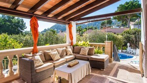 Un espace extérieur aménagé, abrité, avec un canapé et des fauteuils, une table, et une vue sur une piscine et la verdure.