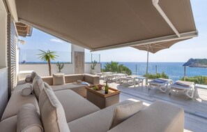 Ein sonnendurchfluteter Außenbereich mit Meerblick, ausgestattet mit einer Couch, Tischen und Sonnenschirmen.