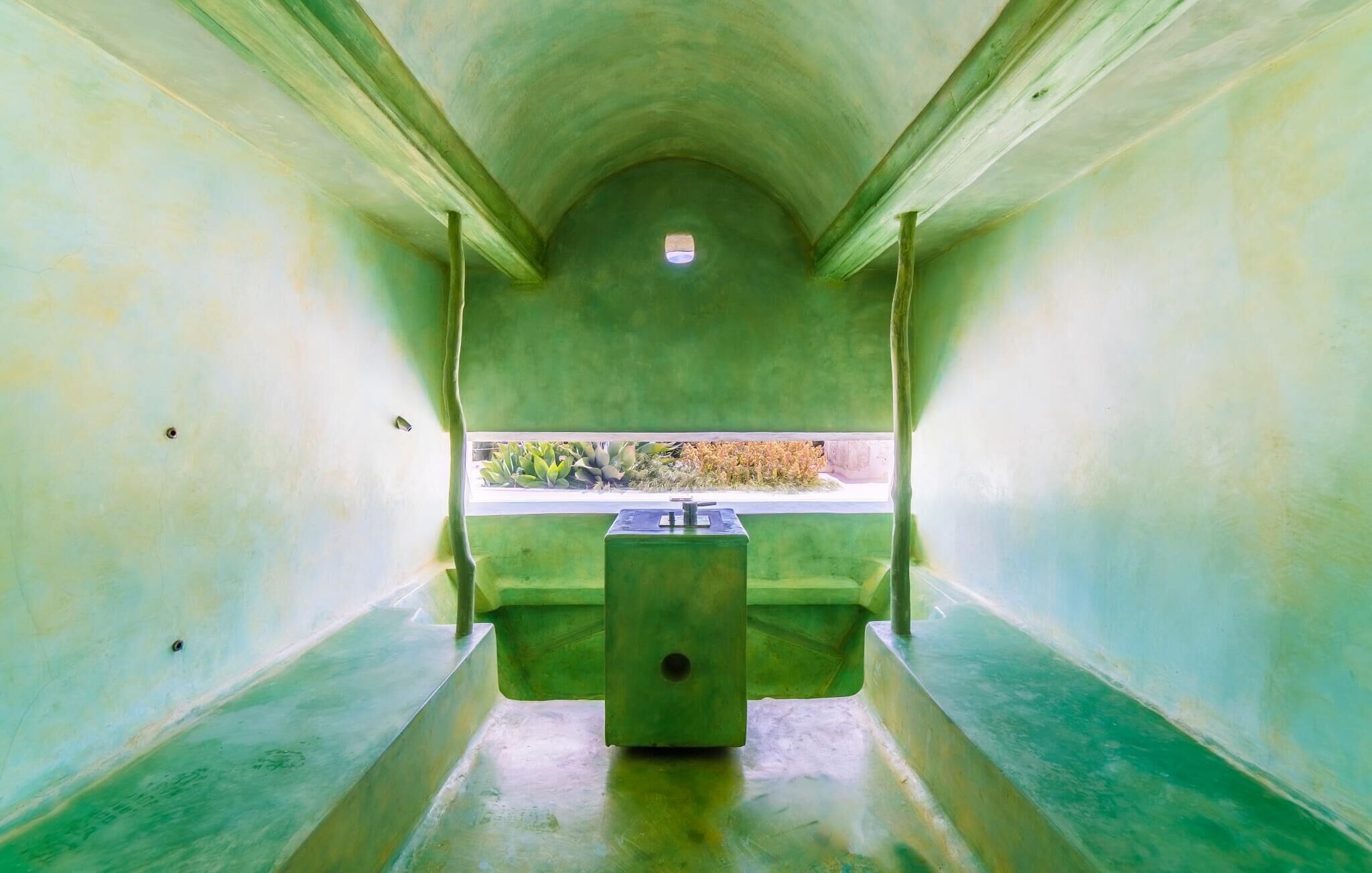 A narrow, green-walled room with a small window at the far end, a bench, and a table with a water dispenser.