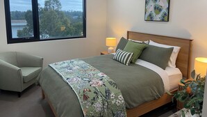 Een slaapkamer met een groot raam, een bed met groene beddengoed, een stoel, een nachtkastje met een lamp en een aan de muur hangend schilderij.