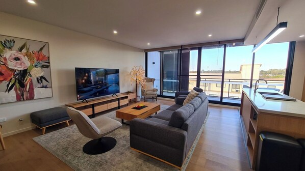 A modern living room with a large flat-screen TV, a sofa, a coffee table, and a dining area with a kitchen counter. - Huntington Apartment - Right in the heart of Rouse Hill Town Centre (Rouse Hill)