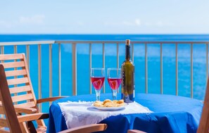 Una mesa con una botella de vino y dos copas de vino tinto, un plato de galletas y una silla de madera en un balcón con vista al mar.