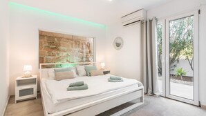 A modern bedroom with a stone accent wall, a large bed with white bedding, two bedside tables with lamps, and a view of greenery through the window. - Beautiful home in Sa Font de Sa Cala (Sa Font de Sa Cala)