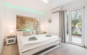 A modern bedroom with a stone accent wall, a large bed with white bedding, two bedside tables with lamps, and a view of greenery through the window. - Beautiful home in Sa Font de Sa Cala (Sa Font de Sa Cala)