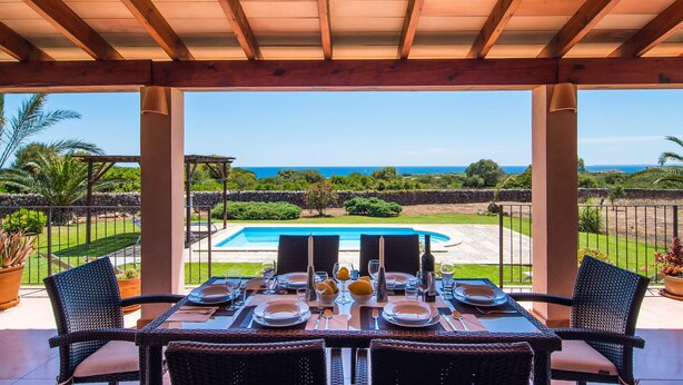 Un área de comedor al aire libre techada con vista al océano, una piscina y un muro de piedra.
