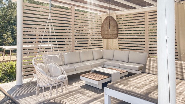 Una zona relax esterna coperta con un divano bianco, una sedia sospesa e un tavolino in legno.