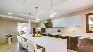 Una cocina moderna con una isla central, taburetes blancos tipo bar, iluminación colgante y vistas al área de comedor.