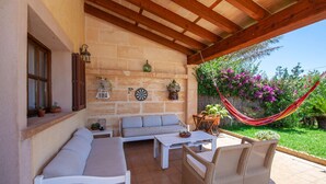 Eine überdachte Terrasse mit einer Steinwand, Holzbalken, einer Couch, einem Tisch, Korbstühlen, einer Hängematte und einem blühenden Garten.