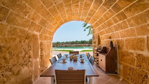 Un espace extérieur aménagé pour les repas, avec une arche en pierre, une table dressée avec des assiettes et des tasses, et une vue sur une piscine et une végétation luxuriante en arrière-plan.