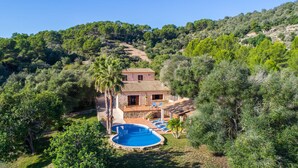Una casa a due piani con piscina, circondata da rigogliosa vegetazione e colline.