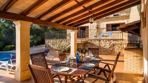 Zona pranzo all'aperto con parete in pietra, sedie di legno e tavolo imbandito per due persone.