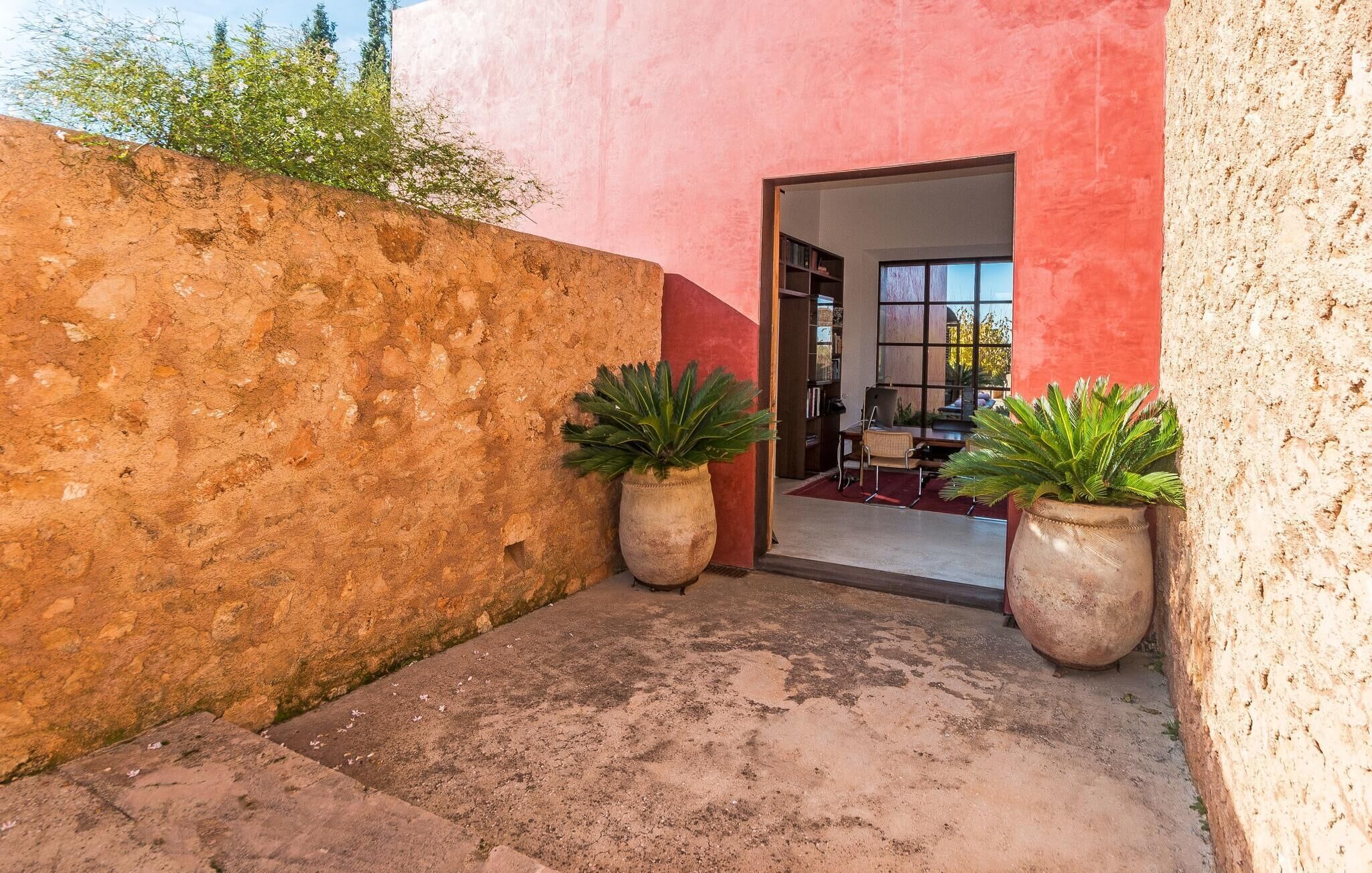 Un estrecho pasillo exterior con una pared roja y una puerta que da a una habitación con un ventanal grande. Hay dos plantas en macetas a ambos lados del pasillo.