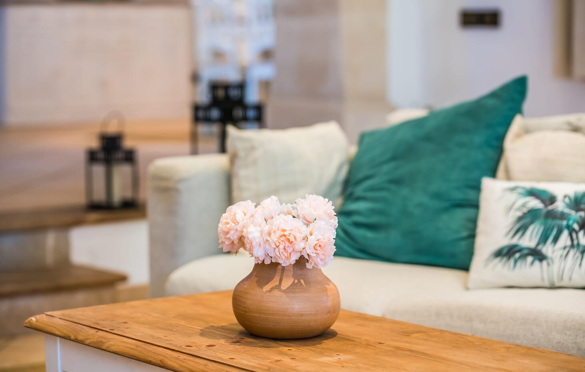 A wooden table with a vase of flowers in a room with a beige sofa, a green pillow, and a palm print pillow.