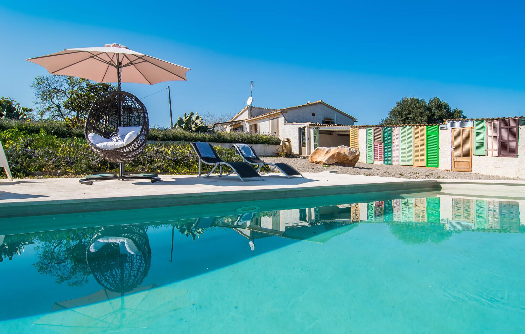 Una piscina con lettini da sole e una sedia sospesa, circondata da una casa e un cancello colorato.