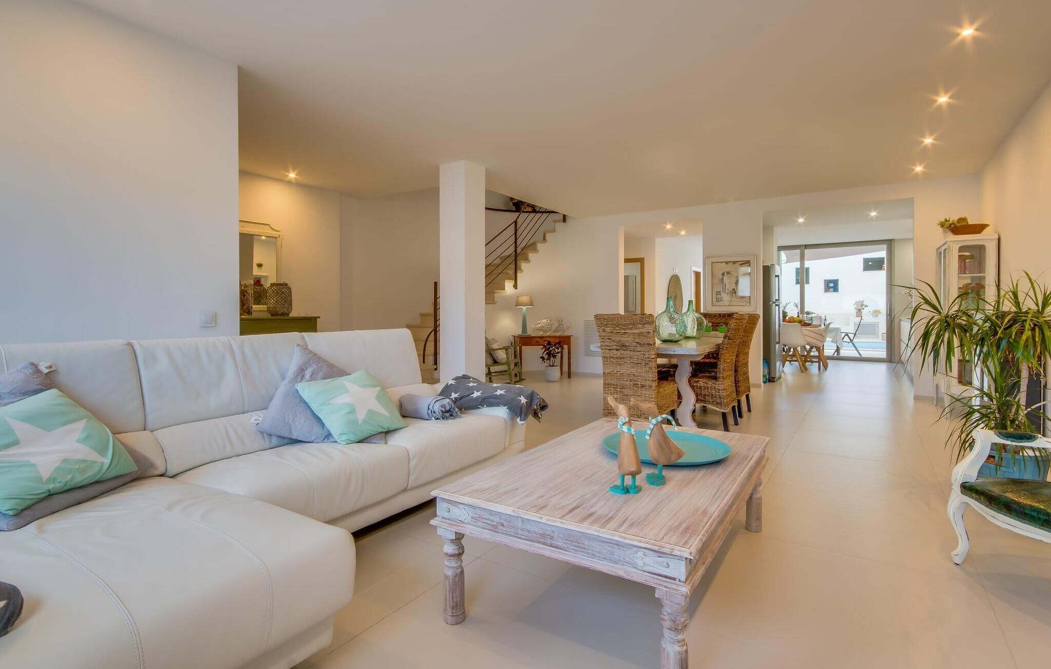 Een ruime woonkamer met een witte bank, een houten salontafel en een eethoek met rieten stoelen.