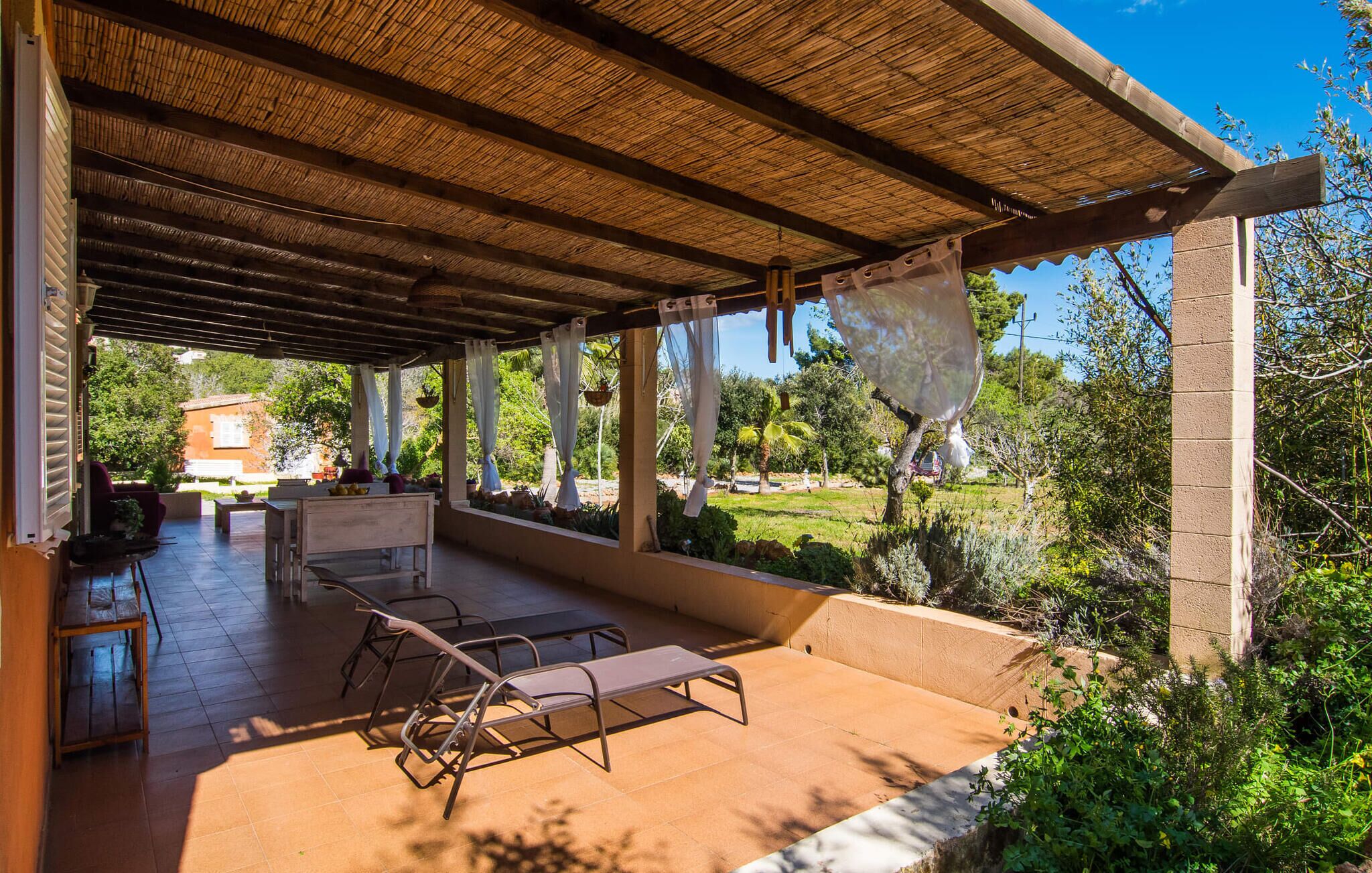 An outdoor covered patio with a thatched roof, a seating area with metal chairs, and a view of a garden and building in the background.