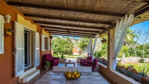 A covered patio with two red chairs, a wooden table with a basket of lemons, and a view of greenery and buildings in the background.