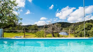 A clear swimming pool with a wooden fence, a blue umbrella, and a view of a mountainous landscape.