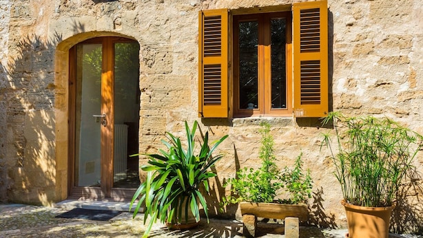 Un bâtiment rustique en pierre, doté d’une porte en bois et de volets aux fenêtres, ainsi que de plantes en pot et d’un banc.