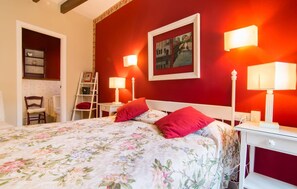 A bedroom with a floral bedspread, two red pillows, a white headboard, and a red accent wall. There are two bedside lamps and a framed picture above the bed. - La Casa de los Espejos (Capdepera)