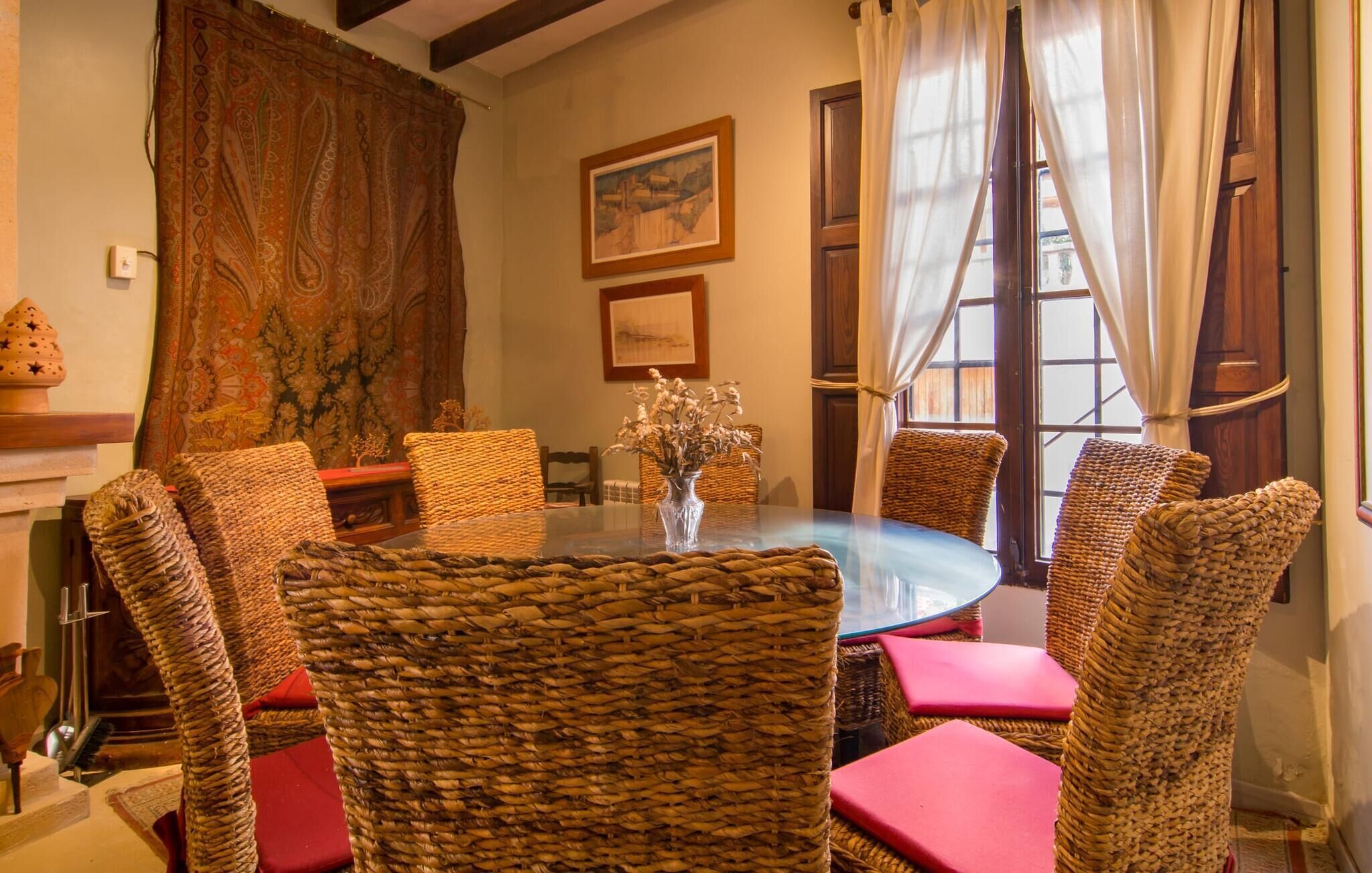 A round table with wicker chairs, a vase with dried flowers, and a painting on the wall.