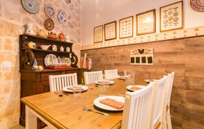 A dining area with a wooden table, white chairs, and a stone wall with decorative plates and framed pictures. - La Casa de los Espejos (Capdepera)