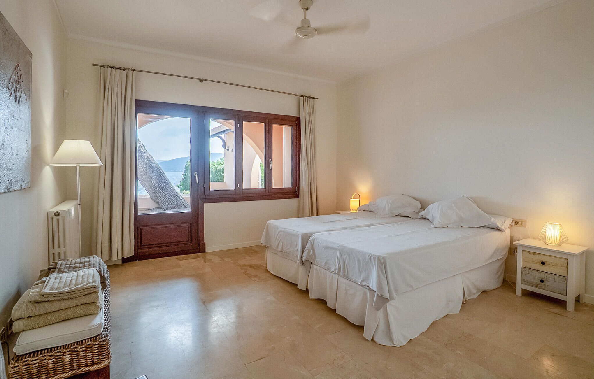 A bedroom with two beds, a large window with curtains, a radiator, a lamp, and a bedside table.