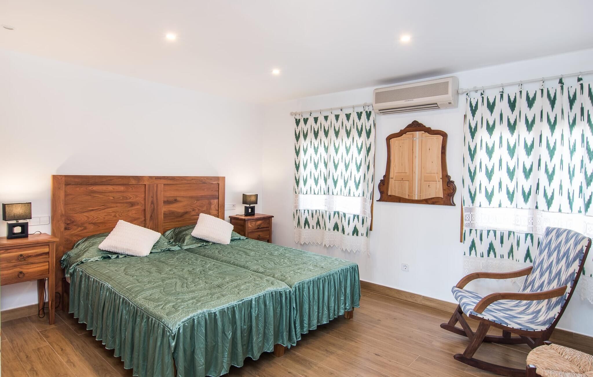 A bedroom with a wooden headboard bed, two bedside tables with lamps, a rocking chair, and patterned curtains.