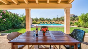 A wooden table with a bottle and glasses, a flower vase, and a view of a swimming pool under a pergola. - Stunning home in Alcudia with private swimming pool, can be inside or outside (Alcudia)