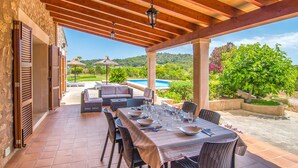 Un área de comedor al aire libre techada con vista a la piscina, mobiliario de mimbre y una mesa dispuesta para seis personas.