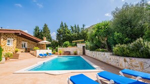 Ein Swimmingpool mit blauem Wasser, umgeben von einer Steinmauer und verschiedenen Pflanzen. Liegestühle und ein kleiner Tisch mit Sonnenschirm sind vorhanden.