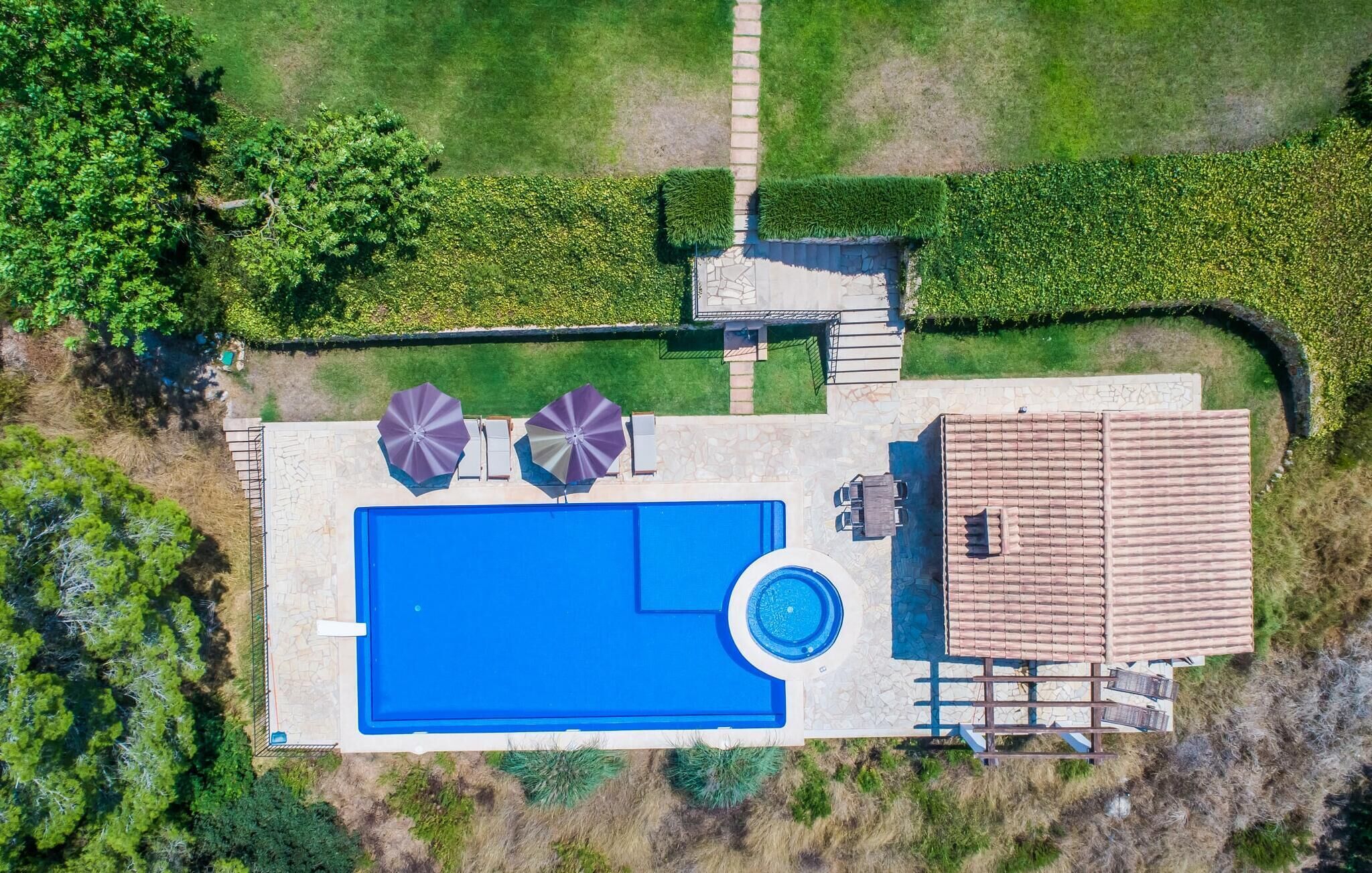 An aerial view of a property with a swimming pool, two umbrellas, a patio area, and a covered patio with a tiled roof.