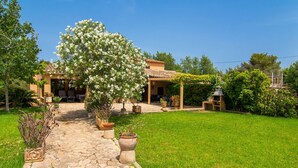 Eine Villa mit einem gepflasterten Weg, der zu einer überdachten Terrasse führt, umgeben von üppiger Vegetation und blühenden Bäumen.