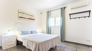Ein Schlafzimmer mit einem Bett, Nachttischen, einem Fenster mit Vorhängen, einer Klimaanlage und einem an der Wand befestigten Regal.