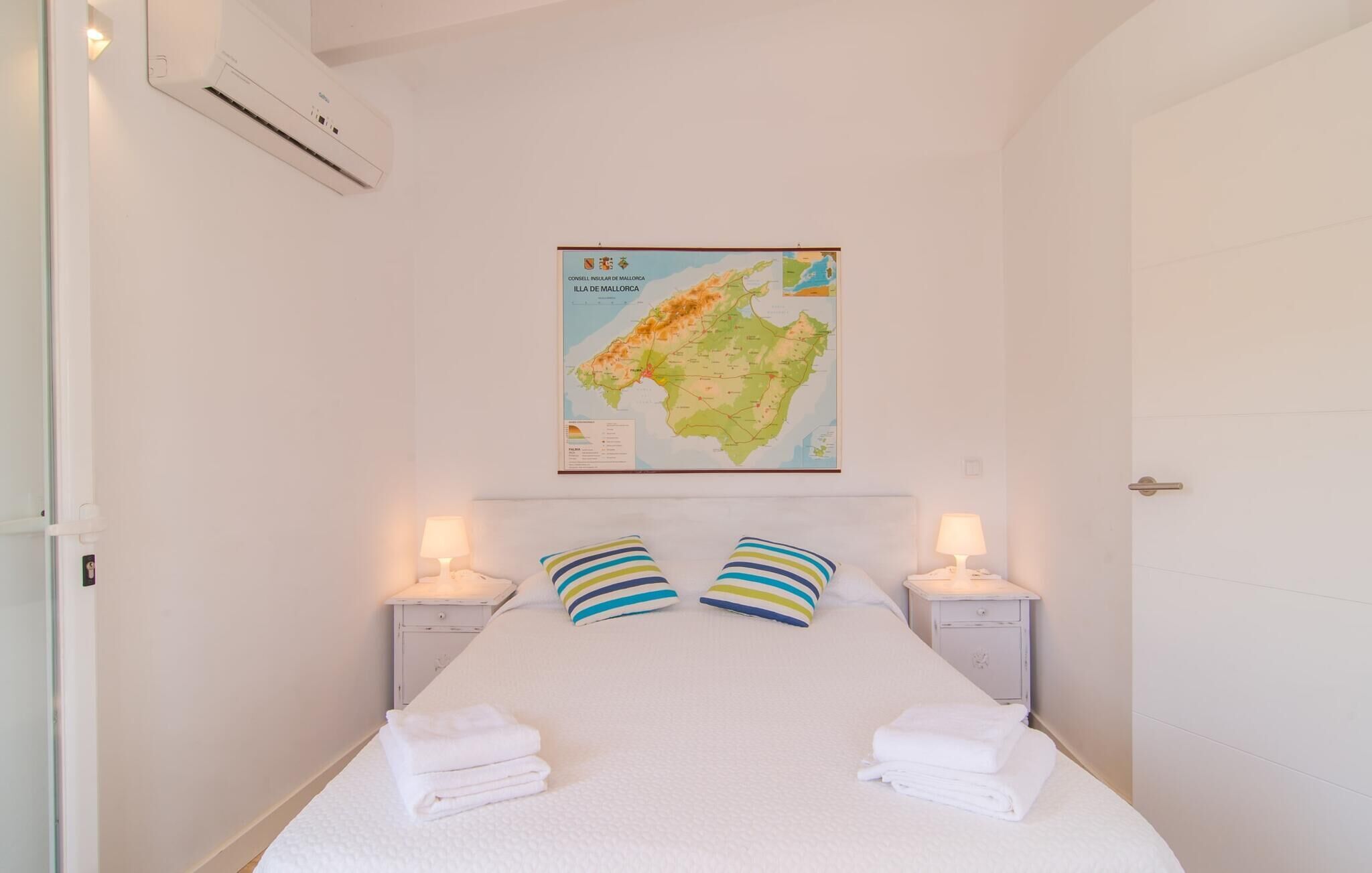 Een net opgemaakt bed met witte lakens, twee gestreepte kussens en gevouwen handdoeken aan weerszijden. Een kaart van Mallorca hangt aan de muur boven het bed.