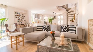 Ein modernes Wohnzimmer mit einem grauen Ecksofa, einem Glastisch und einem Holzstuhl. Im Hintergrund ist eine Küche mit weißen Schränken und einer Wendeltreppe zu sehen.