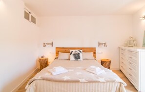 Une chambre à coucher avec une tête de lit en bois, deux lampes de chevet, un lit recouvert d’une couverture à motifs d’étoiles, un coussin en forme de carlin et une commode avec des tiroirs.