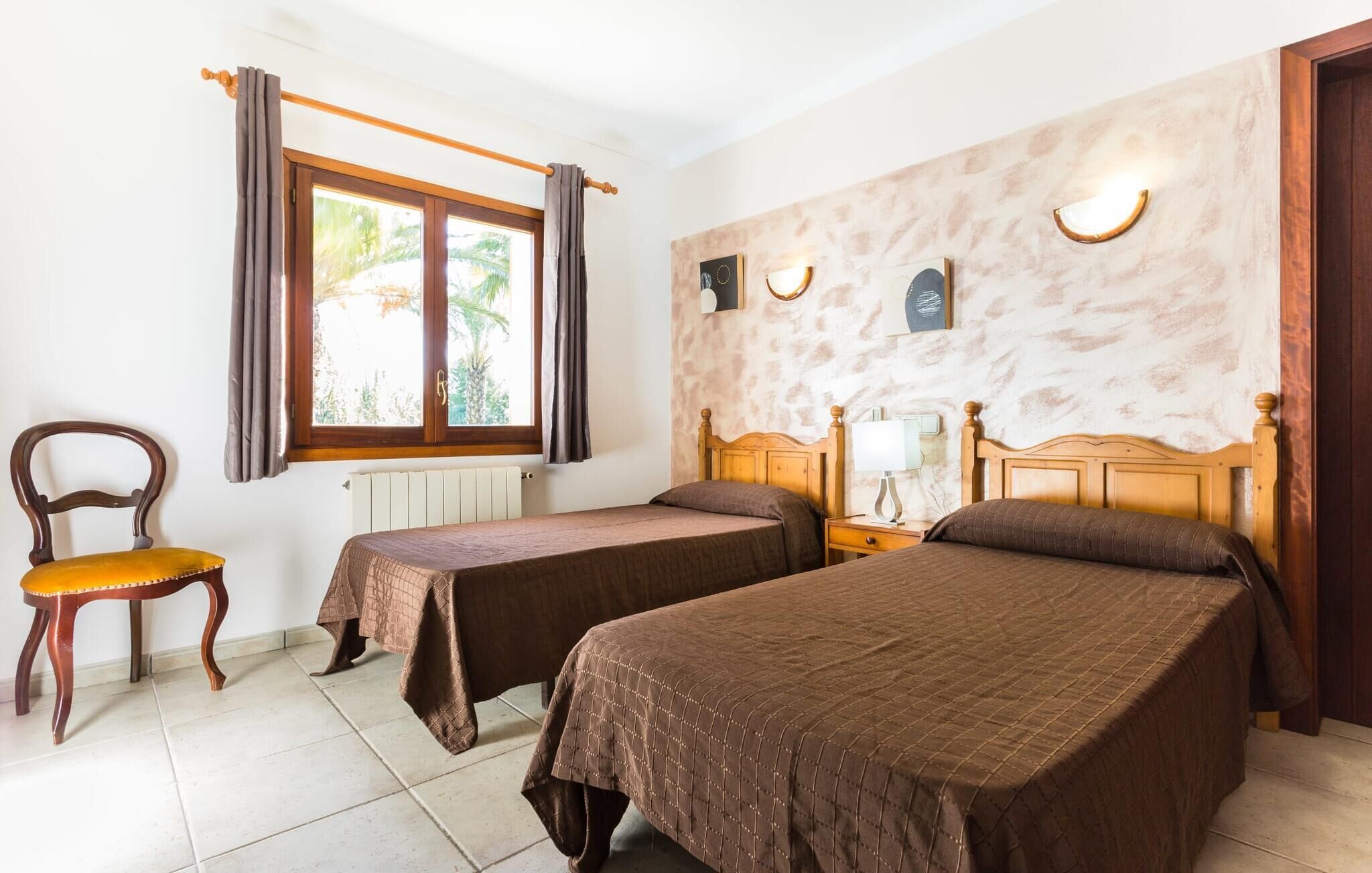 A room with two single beds, a wooden chair, a window with curtains, a radiator, and a wall with a textured finish and wall-mounted lights.