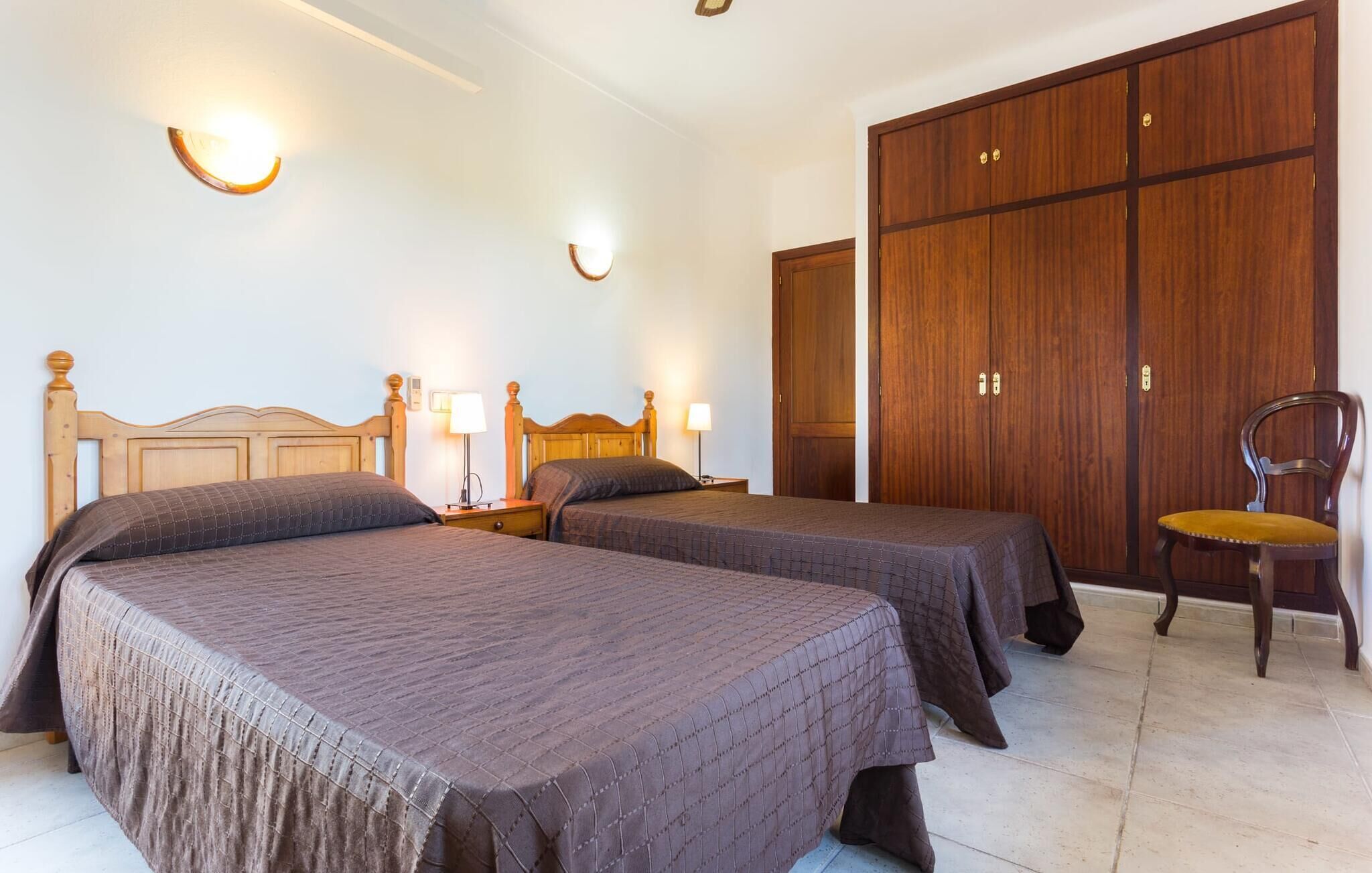 A hotel room with two beds, wooden headboards, bedside lamps, a wardrobe, and a chair.