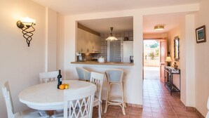 Un comedor con una mesa redonda y cuatro sillas, una cocina con armarios y una lámpara colgante, y un pasillo que conduce a una puerta.