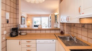 Cocina compacta con lavavajillas, un lavamanos y cafetera. Hay una ventana que proporciona luz natural y un aplique decorativo sobre la encimera.