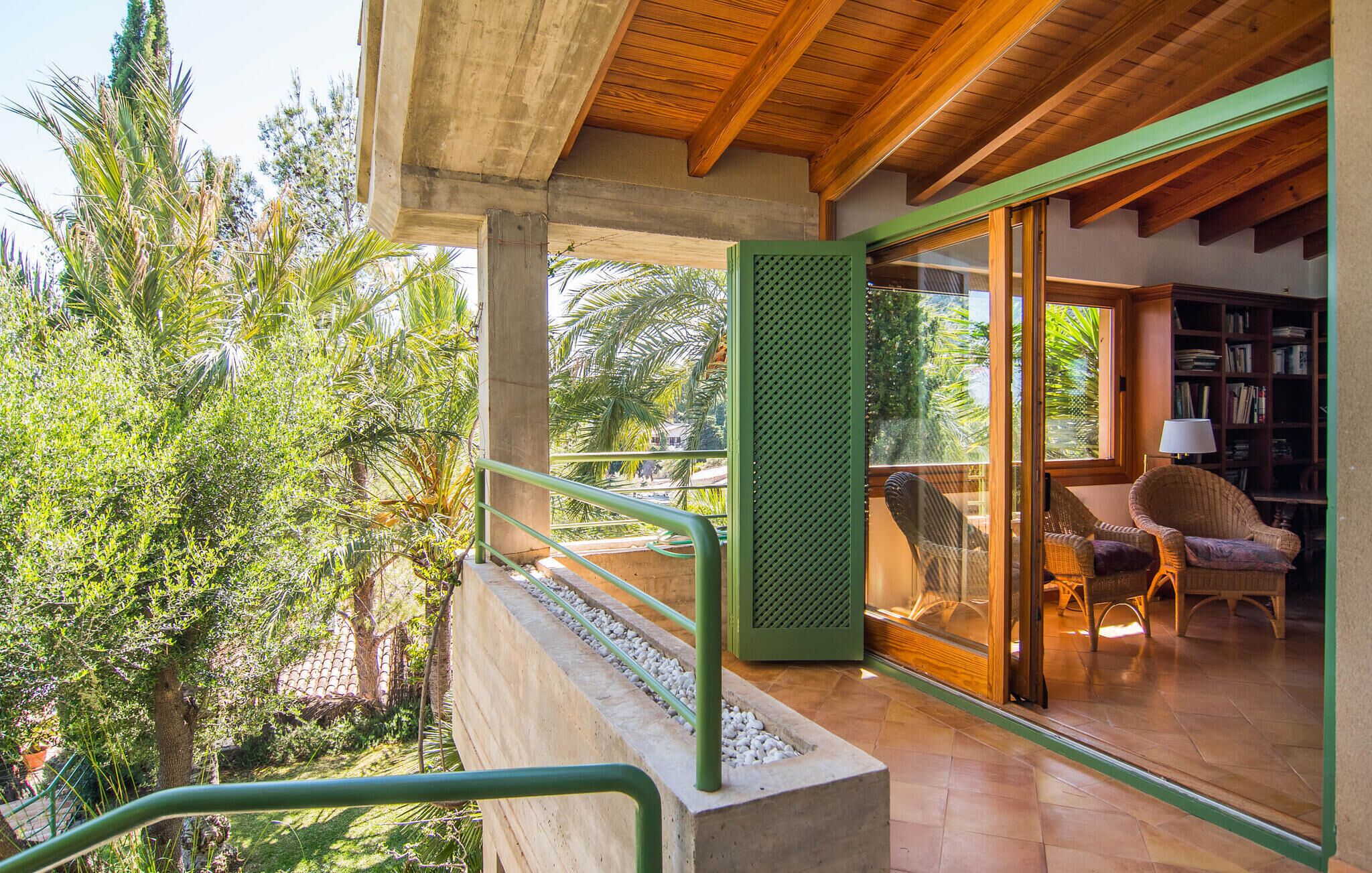 Un balcón con piso de concreto, barandal verde y vista a árboles y una casa. Adentro, hay una silla de mimbre y una estantería.