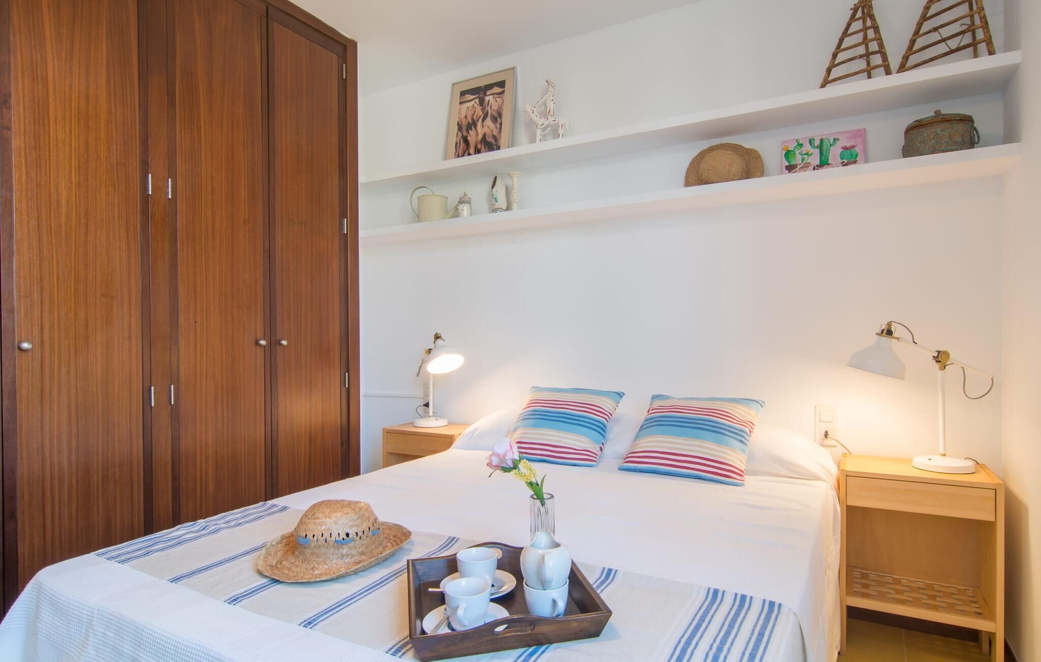 A neatly arranged bedroom with a large bed, bedside tables with lamps, a wooden wardrobe, and decorative items on shelves.