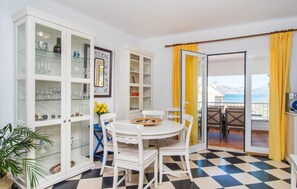 Uma área de refeições com chão xadrez, mobiliário branco e vista para o mar através de uma porta de vidro.