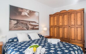 Quarto de hotel com uma cama grande, um guarda-roupa de madeira, um quadro na parede com uma praia e uma mesa de cabeceira com uma luminária.