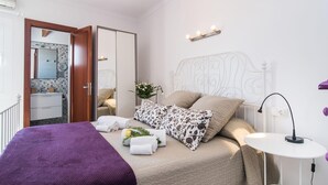Un dormitorio con cama, mesita de noche, lámpara, espejo y baño visible a través de una puerta abierta.