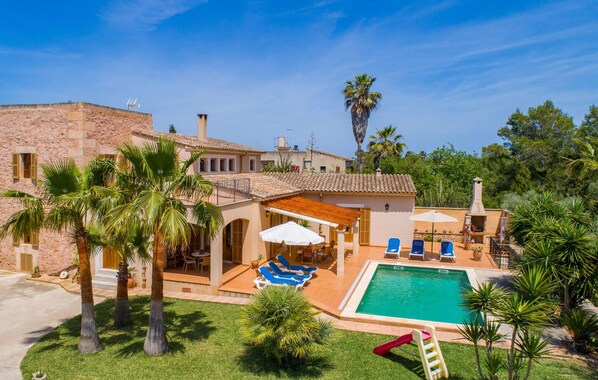 Una villa de estilo mediterráneo con piscina, mobiliario exterior y exuberante vegetación.