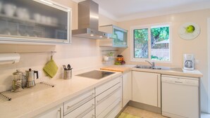 A modern kitchen with white cabinets, a stainless steel range hood, a built-in oven, a coffee maker, and a window with a view of greenery.