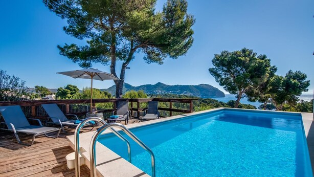 A pool with clear blue water, surrounded by wooden deck chairs and umbrellas, overlooking a scenic view of mountains and the sea. - Stunning home in Canyamel with private swimming pool, can be inside or outside (Canyamel)
